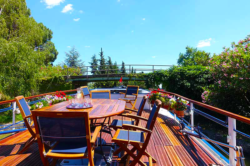 La soleada cubierta del Anjodi con uno de los puentes peatonales del Canal du Midi al fonto... ANJODI CRUCEROS FLUVIALES CANALES DE FRANCIA EUROPEAN WATERWAYS BARGE CRUISES LUXURY BARGE CRUISES ANJODI CRUCEROS EN BARCO PEQUEÑO CRUCEROS CANAL DU MIDI CRUCEROS FAMILIA CRUCEROS FAMILIARES CRUCEROS DE LUJO CRUCEROS DIFERENTES CRUCEROS POCOS PASAJEROS CRUCEROS LUXURY CRUISES CRUCEROS GASTRONOMIA CRUCEROS CHEF